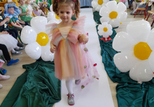 Dziewczynka z grupy "Motylki" w kolorowej sukience i w welonie w kwiatki idzie po białym dywanie. Dziewczynka prezentuje swoją kreację.