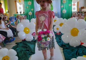 Ula z grupy "Motylki" idzie po białym dywanie. Dziewczynka ubrana jest w różową bluzeczkę, spódniczkę, getry w kwiatki i kapelusz, a ręku trzyma koszyk z kwiatkami.