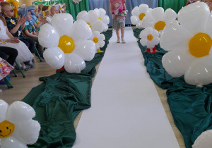Ula z grupy "Pszczółki" idzie po białym dywanie pomiędzy balonami w kształcie stokrotek i prezentuje swoją kreację kwiatową. Dziewczynka trzyma w ręku koszyk z kwiatami. W tle dekoracja z okazji Dnia Ziemi, a po bokach siedzi publiczność.