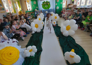 Dziewczynka z grupy "Pszczółki" prezentuje swoją kwiatową kreację idąc po białym dywanie wśród balonów w kształcie stokrotek. Ubrana jest w białą sukienkę w kwiatki, a na głowie ma kaopelusz. Po bokach sali siedzą na krzesełkach przedszkolaki ze wszystkich grup. W tle dekoracja z okazji Dnia Ziemi.
