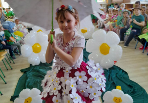 Oliwia z grupy "Słoneczka" prezentuje swoją kreację podczas pokazu mody. Dziewczynka ubrana jest w kwiatową spódnicę i bluzkę. Na głowie ma wianuszek, a w rękach trzyma rozłożoną kwiatową parasolkę.