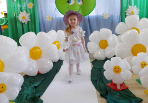 Dziewczynka w białej sukience w kwiatki i w kapeluszu na głowie idzie po białym dywanie wśród balonów w kształcie stokrotek. W tle dekoracja z okazji Dnia Ziemi.