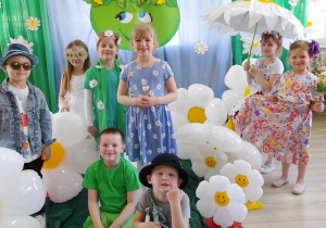 Ośmioro dzieci z grupy "Słoneczka" stoi pomiędzy balonami w kształcie stokrotek i pozuje do zdjęcia w strojach kwiatowych. W tle dekoracja z okazji Dnia Ziemi.