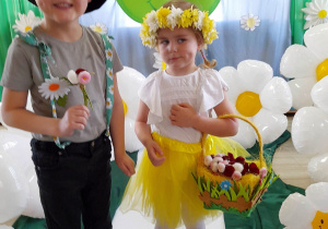 Dwoje dzieci stoi na białym dywanie wśród stokrotkowych balonów. Tomek z grupy "Słoneczka" trzyma bukiecik stokrotek w ręku, a dziewczynka z grupy "Pszczółki" trzyma koszyk z kwiatkami.