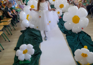 Vanessa z grupy "Słoneczka" idzie po białym dywanie w białej sukni z kwiatkami i stokrotkowych okularach. Po bokach siedzą dzieci - publiczność. W tle dekoracja z okazji Dnia Ziemi.