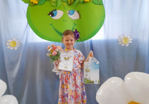 Gabrysia w kwiatowym stroju stoi na tle dekoracji z okazji Dnia Ziemi. Dziewczynka trzyma w rękach drobne upominki i dyplom.