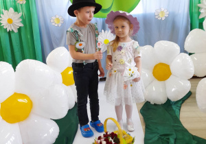 Dwoje dzieci ubranych w kwiatowe stroje stoi na białym dywanie na tle dekoracji z Okazji Dnia Ziemi. Przed nimi znajduje się koszyk ze stokrotkami. Chłopiec podaje dziewczynce bukiecik stokrotek.