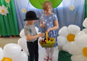 Tomek wraz z Alicją z grupy "Słoneczka" stoją na białym dywanie. Dziewczynka ma na sobie niebieską sukienkę w kwiatki i trzyma koszyk ze stokrotkami, a chłopiec wyjmuje z koszyka kwiatki. W tle dekoracja z okazji Dnia Ziemi, a obok dzieci stoją stokrotkowe balony.