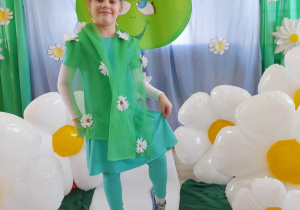 Alicja z grupy "Słoneczka" prezentuje się wśród balonów w kształcie stokrotek i na tle dekoracji z okazji Dnia Ziemi. Dziewczynka ubrana jest w zielone getry, sukienkę oraz szal w stokrotki, a na głowie ma kwiatkowy wianek.