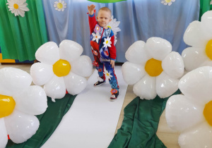 Chłopiec z grupy "Motylki" pozuje do zdjęcia w kwiatowym stroju. W tle dekoracja z okazji Dnia Ziemi, a obok chłopca stoją balony w kształcie stokrotek.