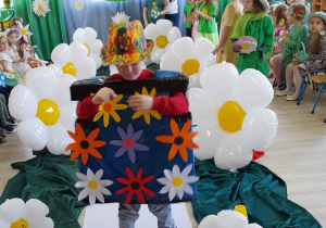 Bartek z grupy "Biedronek" w stroju kwiatowego robota idzie po białym dywanie wśród stokrotkowych balonów.