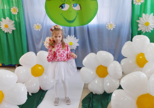 Dziewczynka z grupy "Motylki" ubrana w bluzkę w kwiatki, białą spódniczkę oraz wianuszek na głowie pozuje do zdjęcia na tle dekoracji z okazji Dnia Ziemi.