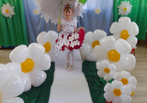 Oliwia pozuje do zdjęcia na tle dekoracji z okazji Dnia Ziemi. Dziewczynka ubrana jest w spódniczkę i bluzkę w kwiatki, na głowie ma wianek, a w ręku trzyma parasol.