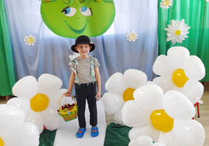 Tomek z grupy "Słoneczka" pozuje do zdjęcia na tle dekoracji z okazji Dnia Ziemi. Chłopiec ma spodnie z kwiatowymi szelkami i kapelusz na głowie, a w reku trzyma koszyk ze stokrotkami.