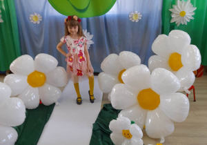 Zosia w kwiatowej sukience prezentuje się na białym dywanie na tle dekoracji z okazji Dnia Ziemi.