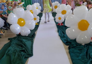 Dziewczynka z grupy "Motylki" ubrana jasnozieloną sukienkę w kwiatki oraz wianek prezentuje się na białym dywanie podczas pokazu mody kwiatowej.
