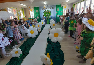 Dzieci ze wszystkich grup stoją po obu stronach sali i składają przyrzeczenie na "Przyjaciela Ziemi". Wzdłuż sali leży biały dywan oraz zielony materiał, na którym stoją balonowe stokrotki. Troje dzieci stoi na tle dekoracji i czyta treść przyrzeczenia.