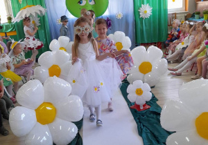 Dzieci z grupy "Słoneczka" idą po białym dywanie prezentując kwiatowe kreacje. Przedszkolaków prowadzi Vanessa w białej sukni z przypiętymi kwiatkami i w stokrotkowych okularach.