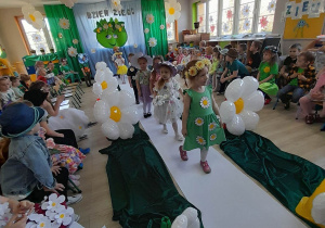 Dzieci z grupy "Pszczółki" idą po białym dywanie podczas pokazu mody. Po bokach sali siedzą koledzy z innych grup.