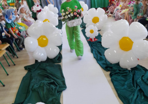 Krzyś z grupy "Słoneczka" idzie po białym dywanie wśród balonowych stokrotek. Chłopiec ubrany jest w zieloną koszulkę i zielone spodnie, a w ręku niesie koszyk z kwiatkami.