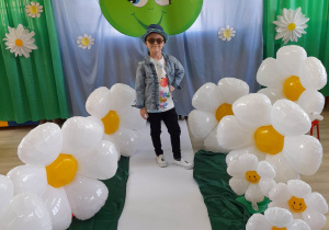 Oskar z grupy "Słoneczka" stoi na białym dywanie. Po bokach na zielonym materiale stoją białe balony w kształcie stokrotek. W tle dekoracja z okazji Dnia Ziemi.