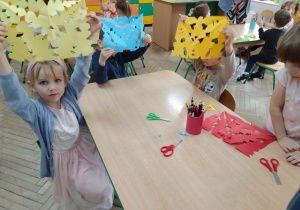 Ula, Mikołaj i Natalia prezentują wykonane przez siebie wycinanki z kolorowego papieru. Na stole leżą nożyczki i kredki.
