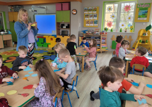 Przedszkolaki siedzą przy stolikach, na których leżą wycięte kawałki papieru kolorowego. Dzieci wykonują kwiaty zgodnie z instrukcją pani z biblioteki.
