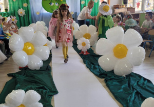 Biedronki w trakcie pokazu mody idą po białym dywanie. Jako pierwsza idzie Zosia w kwiatowej sukience, pozostałe dzieci czekają na swoją kolej