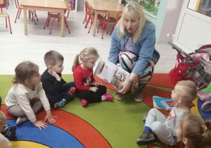 Dzieci siedzą w kole na dywanie i oglądają ilustracje w książce, trzymanej przez panią z biblioteki.