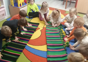 Przedszkolaki siedzą na dywanie i oglądają rozłożone tkaniny łowickie w pasy.