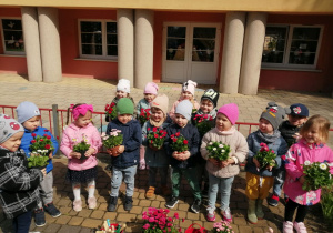 Zdjęcie grupowe dzieci przed budynkiem przedszkola. Dzieci trzymają doniczki ze stokrotkami.