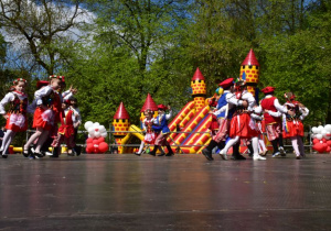 Dzieci w parach tańczą po kole Krakowiaka. W tle przypięte przy scenie biało-czerwone balony, drzewa, dmuchana zjeżdżalnia.