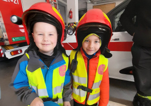Bruno i Oliwier stoją ubrani w hełmy strażackie. Dzieci mają na sobie kamizelki odblaskowe.