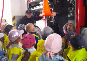 Dzieci stoją przed wozem strażackim, a dwóch strażaków omawia jego wyposażenie. Jeden strażak trzyma w rękach pachołek odblaskowy.