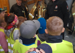 Przedszkolaki podczas wizyty w Straży Pożarnej oglądają powieszone na wieszakach stroje strażackie. Przed dziećmi stoi dwóch strażaków.