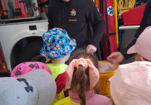 Dzieci stoją w budynku przed wozem strażackim, a strażacy pokazują przedszkolakom sorbent.