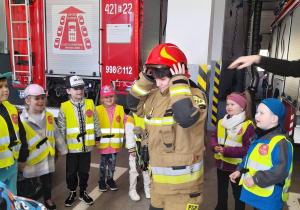 Przedszkolaki stoją w półkolu w budynku Straży Pożarnej, a przed dziećmi pani Ania ubrana w strój strażacki. W tle samochód strażacki.