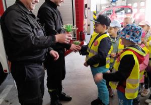 Dwóch chłopców wręcza strażakom wykonane przez dzieci bukiety kwiatków.