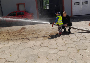 Tomek stoi obok strażaka. Chłopiec trzyma w rękach wąż strażacki i leje wodę w kierunku sztucznego ognia.
