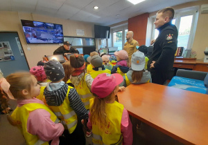 Dzieci oglądają stanowisko kierowania, w którym odbierane są zgłoszenia o pożarach lub wypadkach. Obok stoi trzech strażaków.