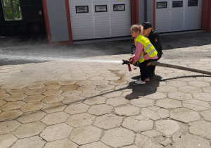Vanessa stoi obok strażaka i leje wodę z węża w kierunku sztucznego ognia.