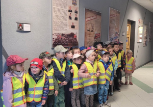 Przedszkolaki stoją ustawione przy ścianie. Dzieci słuchają wykładu panów strażaków na temat bezpieczeństwa.