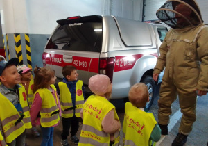 Grupa dzieci w kamizelkach odblaskowych obserwuje strażaka ubranego w kombinezon ochronny na osy i szerszenie. Obok stoi auto z napisem "straż".