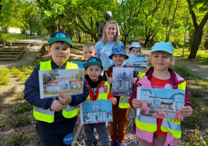 Dzieci stoją razem z panią pod pomnikiem Bolesława Wituszyńskiego w parku. Przedszkolaki trzymają w rękach zdjęcia Kutna.