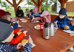 Dzieci siedzą przy stole i degustują kiełbaski upieczone przy ognisku.