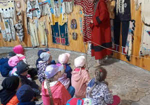 Dzieci siedzą w ziemiance - muzeum i słuchają opowieści przewodnika. W tle stroje indiańskie.