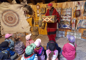 Dzieci siedzą w ziemiance - muzeum, a przewodnik ubrany w strój indiański prezentuje przenośną szafę.