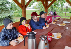 Grupka dzieci siedzi przy drewnianym stole i degustuje kiełbaski.