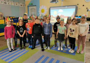 Dzieci stoją w sali przedszkolnej przed tablicą multimedialną. Na głowach mają kolorowe pióropusze indiańskie.