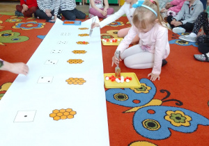 Ula i Oliwia układają pomponiki na "plastrach miodu" według podanej ilości kropek na kartoniku. Wokół siedzą "Motylki" i rodzice i obserwują działania koleżanek.